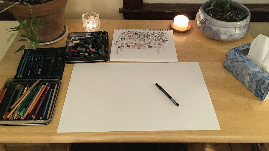 A desk laid out with candles, potted plants, tissues, and art supplies. A blank poster board is front and center, with a smaller sketch pad above showing a table full of food with several dozen stick figures gathered around it.